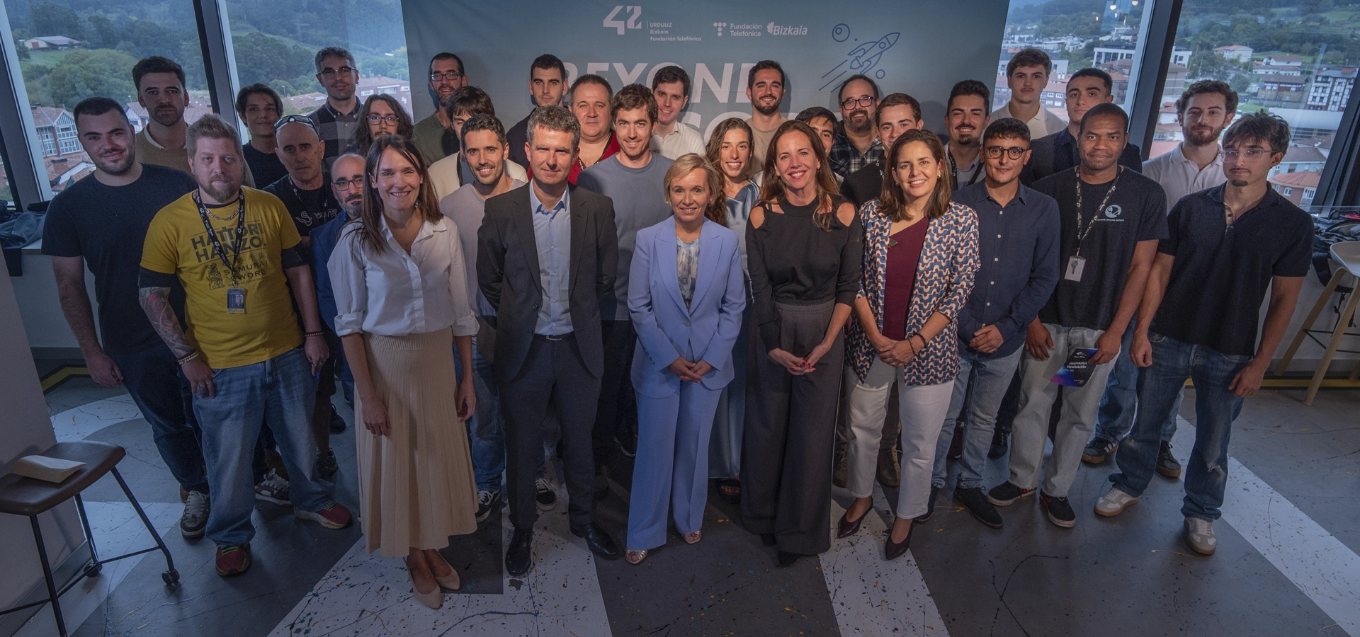 2026ko graduazioa Group of about 30 professionals posing for a group photo in a bright office, with large windows showing a town outside and a blue backdrop with logos in the background.